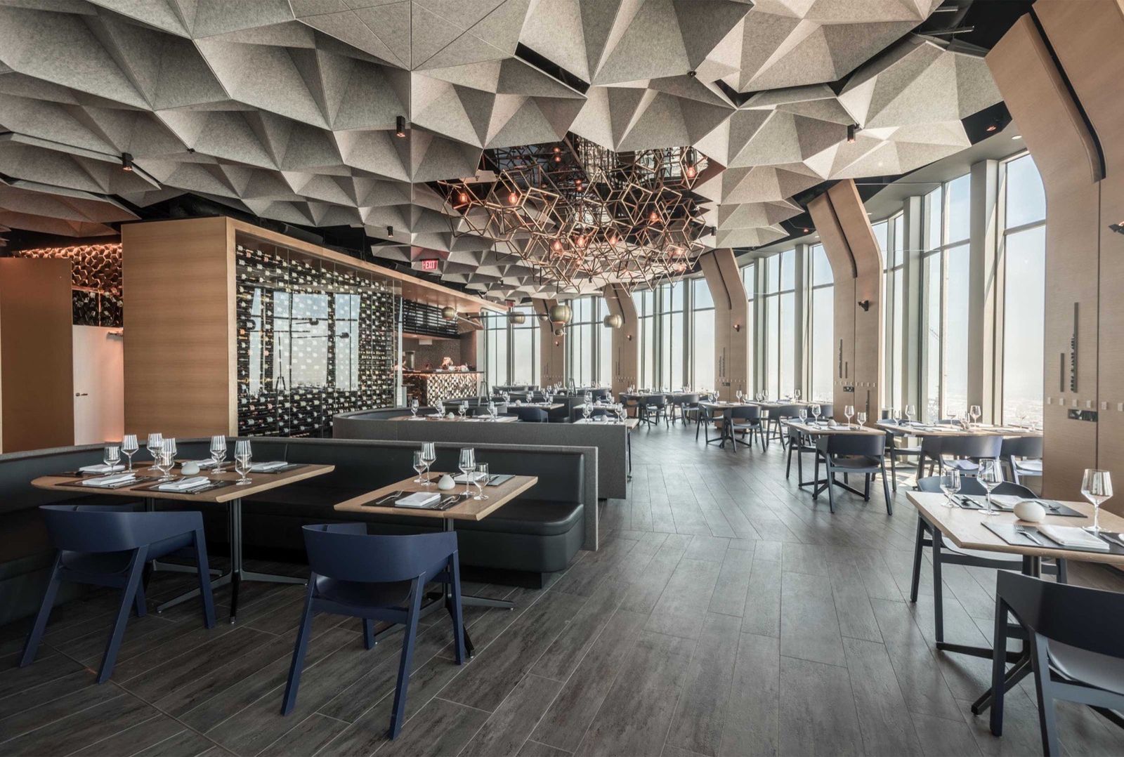 Interior de un restaurante en la última planta: techo de paneles facetados, pared de vinos acristalada y ventanales del suelo al techo sobre la ciudad.